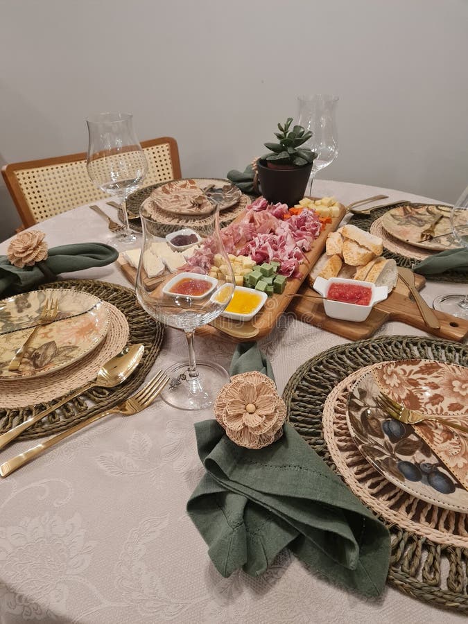 Elegant and rustic table set. A dining table is elegantly set for a meal. It features a light-colored tablecloth and woven placemats. Each setting includes a decorative plate, gold cutlery, and a folded green napkin adorned with a floral napkin ring. Centered is a wooden board with a variety of cheeses, meats, and breads, accompanied by small dishes of condiments. Wine glasses are positioned beside each plate. A small potted succulent adds a touch of greenery to the arrangement.