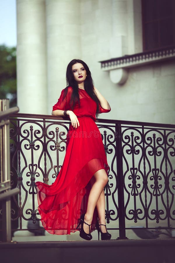 Elegant sensual young woman in red dress posing near a handrail.
