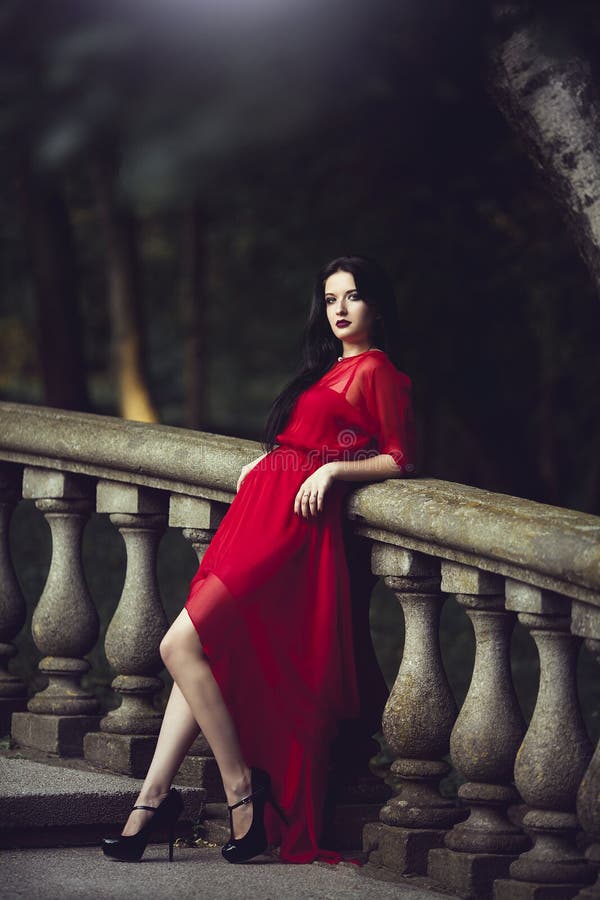 Elegant sensual young woman in red dress posing near a handrail.