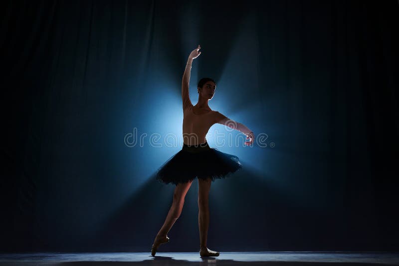 Elegant silhouette of elegant young girl, ballet dancer dancing on stage against dark blue background with spotlight