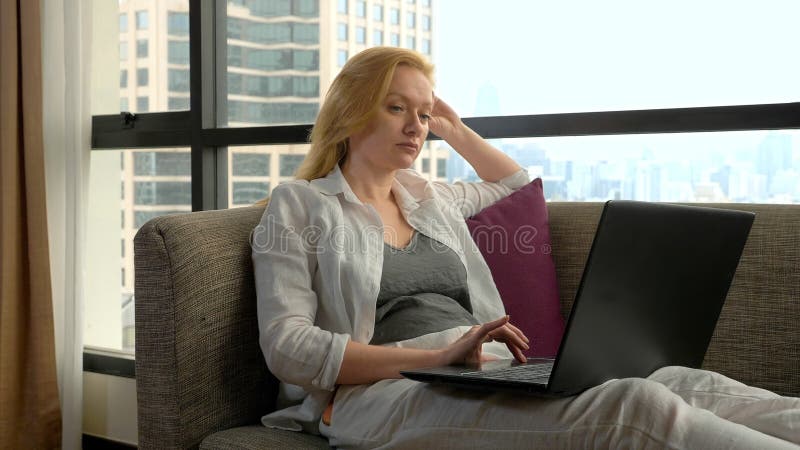 An elegant woman is lying on a sofa by the panoramic window overlooking the skyscrapers and using her laptop royalty free stock image An elegant woman is lying on a sofa by the panoramic window overlooking the skyscrapers and using her laptop royalty free stock image