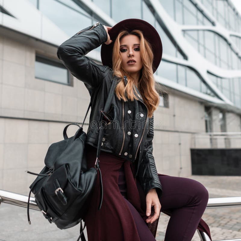 Elegant young woman with red hair in a black jacket with a leather backpack in trendy pants with a cape at the waist sits stock photos Elegant young woman with red hair in a black jacket with a leather backpack in trendy pants with a cape at the waist sits stock photos
