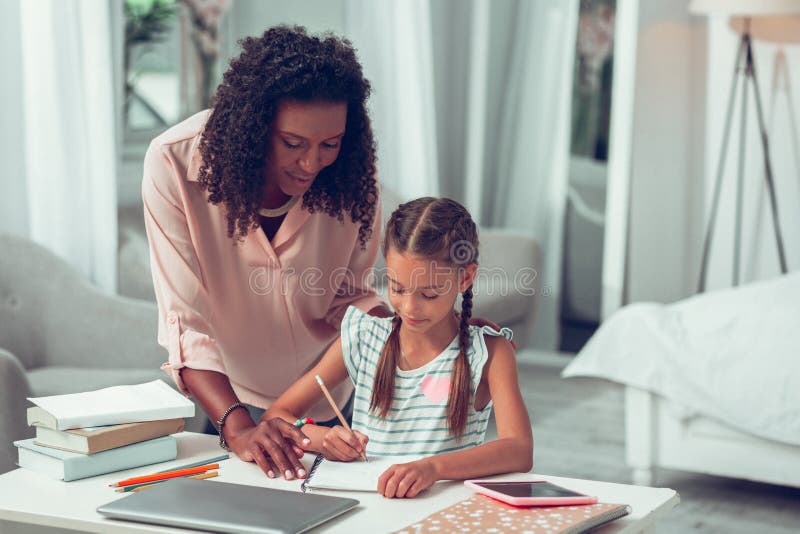 Elegante afroe-amerikanisch Mutter, die ihre Tochter mit dem Handeln einer Hausarbeit unterstützt lizenzfreie stockbilder