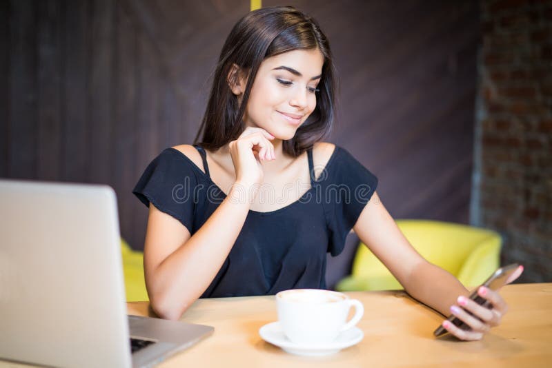 Elegante Jungelächelnfrau mit einer Laptop-Computer an einem Café unter Verwendung eines Handys lizenzfreies stockfoto Elegante Jungelächelnfrau mit einer Laptop-Computer an einem Café unter Verwendung eines Handys lizenzfreies stockfoto