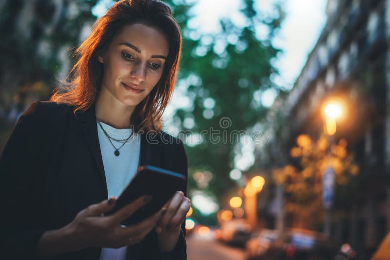 Elegant woman pointing on screen smartphone background lights in night city street, tourist girl using internet technology calls a taxi standing next to road of evening town. Elegant woman pointing on screen smartphone background lights in night city street, tourist girl using internet technology calls a taxi standing next to road of evening town