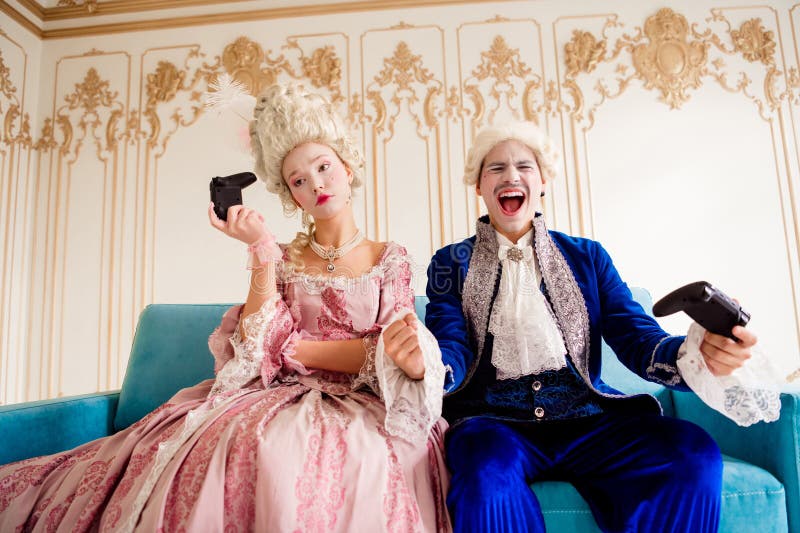 Elegante pareja con elegantes trajes barrocos que disfrutan de juegos modernos en el interior de un palacio histórico. imagen de archivo