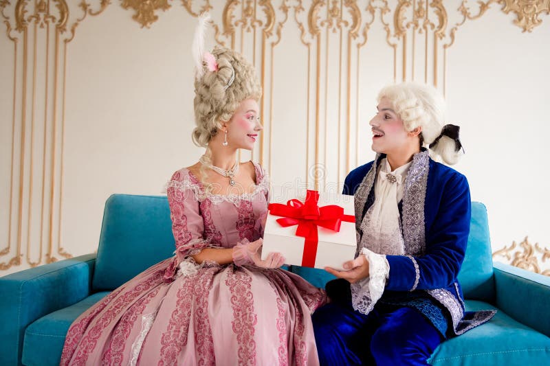 Elegante pareja con trajes barrocos intercambiando un regalo en lujosos interiores fotos de archivo libres de regalías
