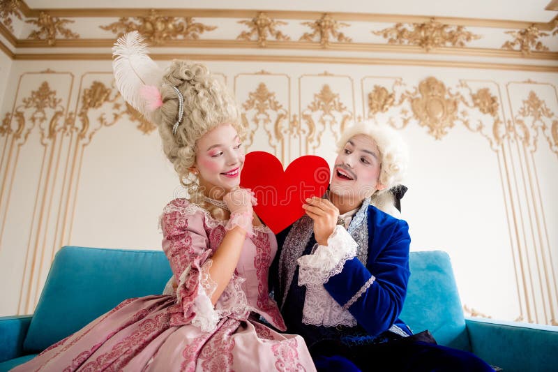 Elegante pareja en trajes temáticos de estilo baroquero sosteniendo un corazón rojo en un interior de palacio histórico imagen de archivo
