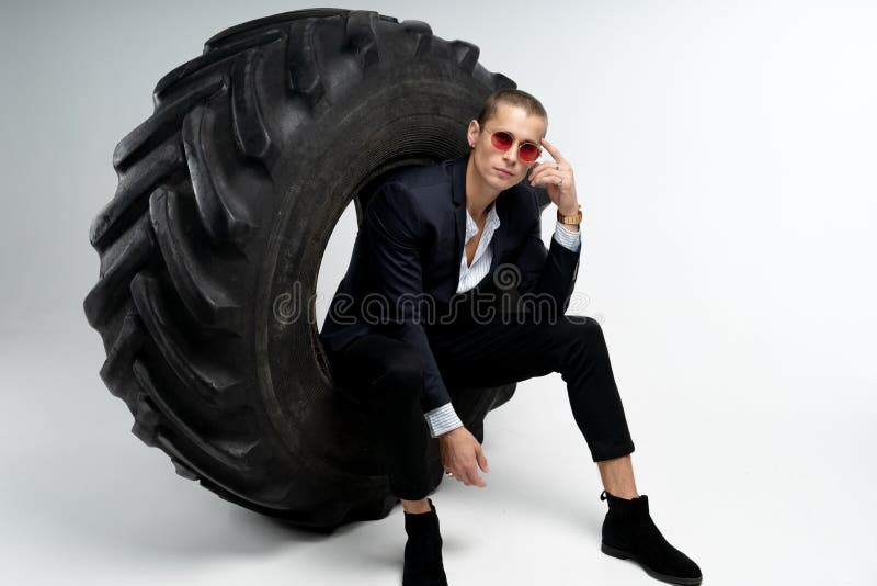 Eleganter junger Gesch?ftsmann im schwarzen Anzug und roten in der Sonnenbrille, die im gro?en Reifen, die Kamera betrachtend sit lizenzfreies stockfoto Eleganter junger Gesch?ftsmann im schwarzen Anzug und roten in der Sonnenbrille, die im gro?en Reifen, die Kamera betrachtend sit lizenzfreies stockfoto