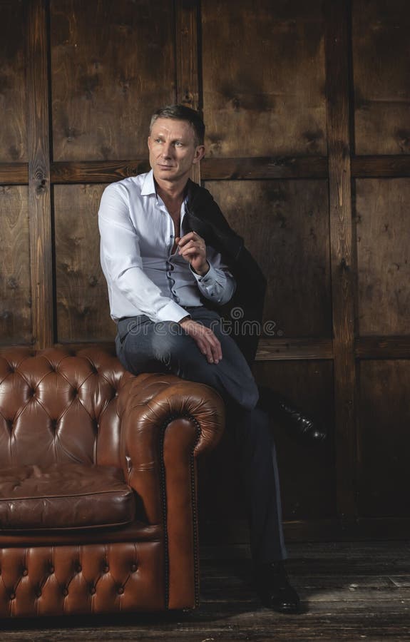 Eleganter moderner Mann in einem Hemd und in einem Mantel sitzt auf einem ledernen Sofa lizenzfreie stockbilder