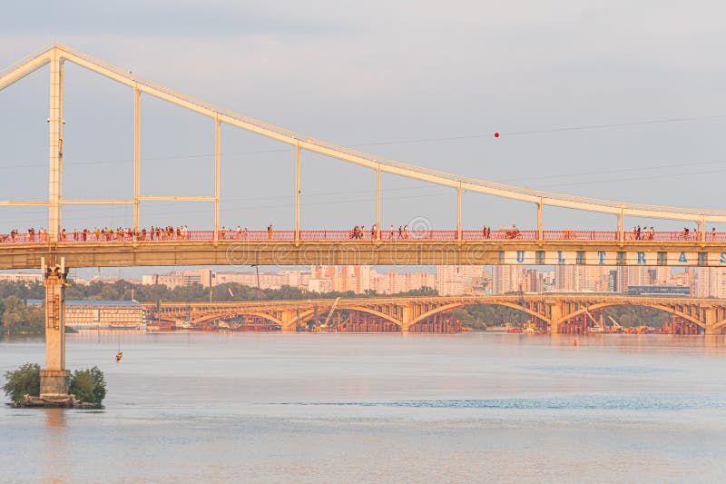 Elevated view of a red suspension bridge with multiple spans over a waterway, supported by sturdy pylons Bridge has cable support structure Warm lighting suggests Kyiv, Ukraine 07-06-2025. Red pylons stock images, royalty-free photos and pictures