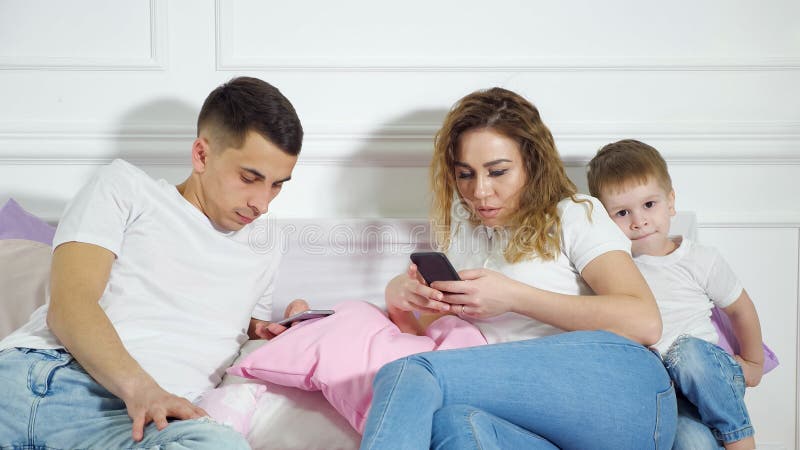 Mom and dad are looking in their mobile phones not paying attention to their child laying in bed. Sad little boy wants. Mom and dad are looking in their mobile phones not paying attention to their child laying in bed. Sad little boy wants