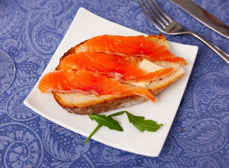 On top of greased bread is slice of lightly salted salmon. Two sandwiches with butter and fish, decorated with arugula leaf. On top of greased bread is slice of lightly salted salmon. Two sandwiches with butter and fish, decorated with arugula leaf