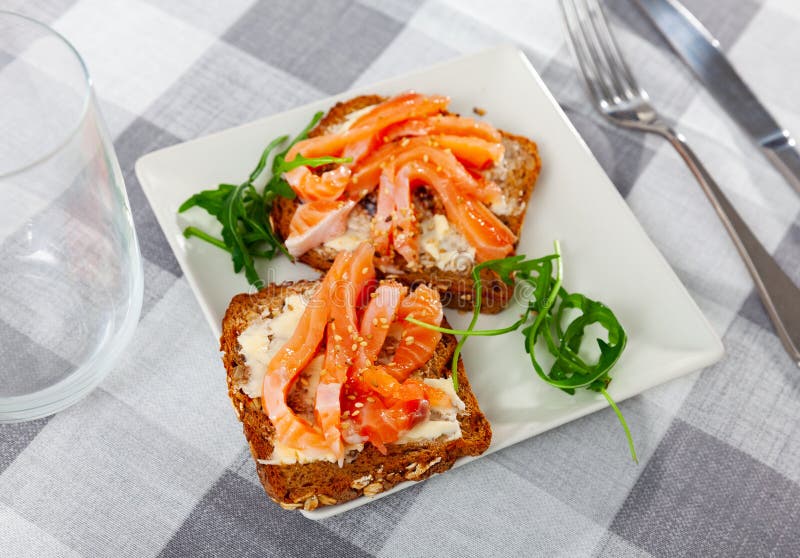 On top of greased bread is slice of lightly salted salmon, sprinkled with sesame seeds. Two sandwiches with butter and fish, decorated with arugula leaf. On top of greased bread is slice of lightly salted salmon, sprinkled with sesame seeds. Two sandwiches with butter and fish, decorated with arugula leaf