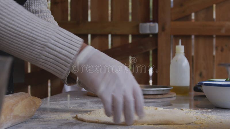 A man in an apron is rolling out pizza dough on a table sprinkled with flour. On the table in front of him are bowls of