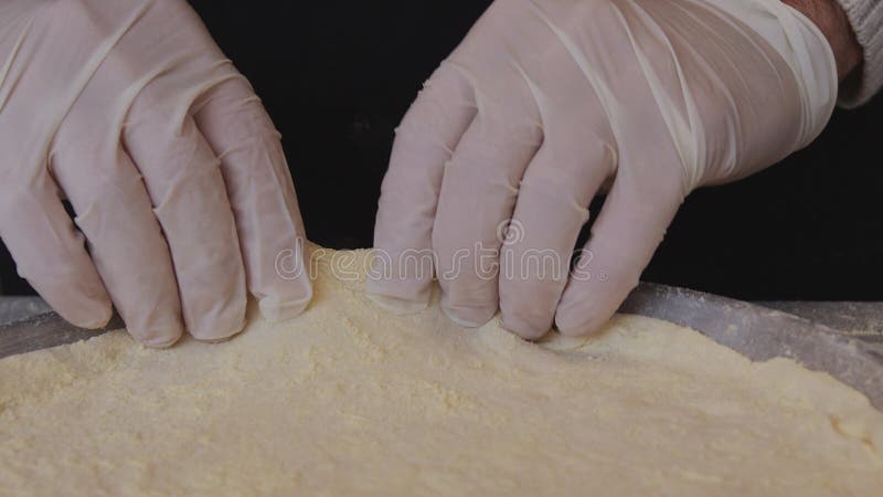 A man in an apron is rolling out pizza dough on a table sprinkled with flour. On the table in front of him are bowls of