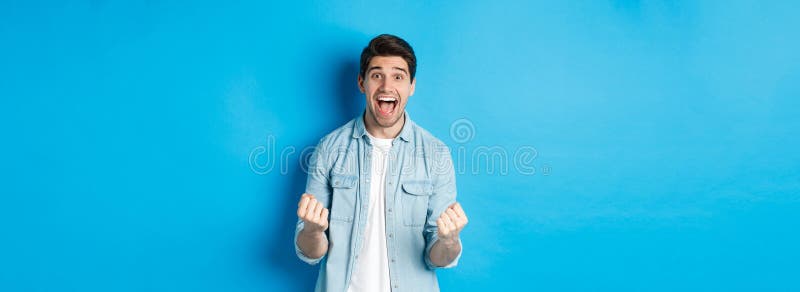 Emocionado hombre apuesto que ganó sintiéndose afortunado y regocijándose de celebrar la victoria y gritando sí contra el azul imagen de archivo