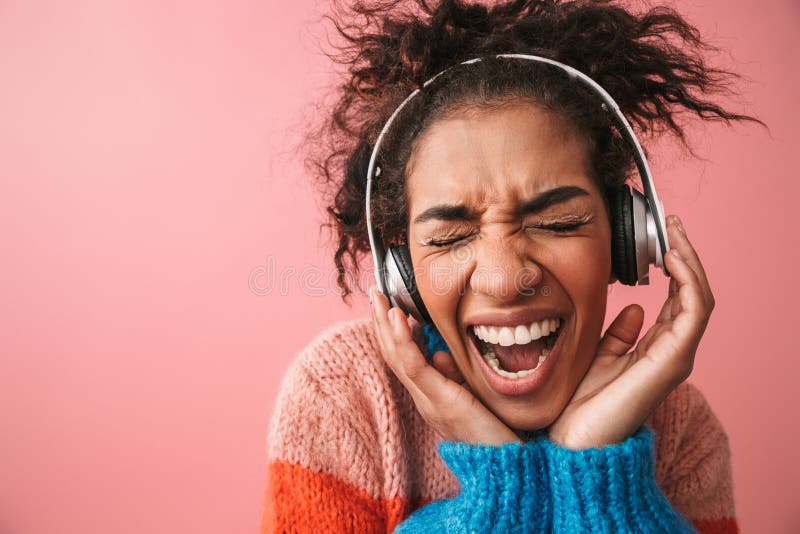 Emotional beautiful young african woman posing  over pink wall background listening music with headphones