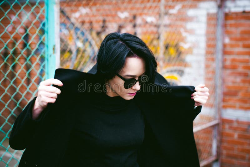 Emotional fashionable portrait of a young brunette woman in black clothes, jeans T-shirt, coat and sunglasses, in a Gothic style s