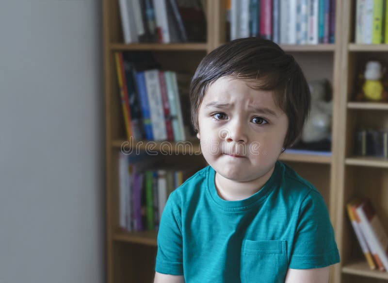 Emotional portrait sad kid looking at camera with unhappy face, Angery little boy sitting alone with blurry bookshelf background