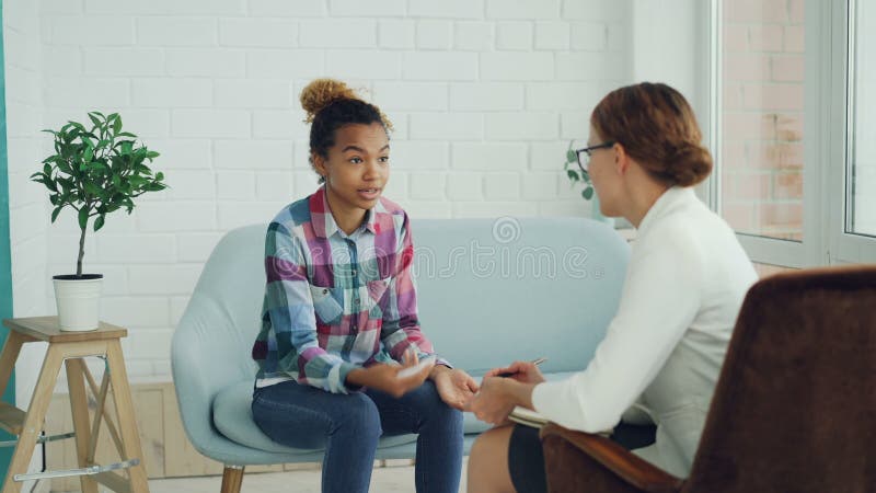 Emotionale Afroamerikanerdame spricht mit dem Psychologen, der auf Couch sitzt, während der Beratung spricht und gestikuliert