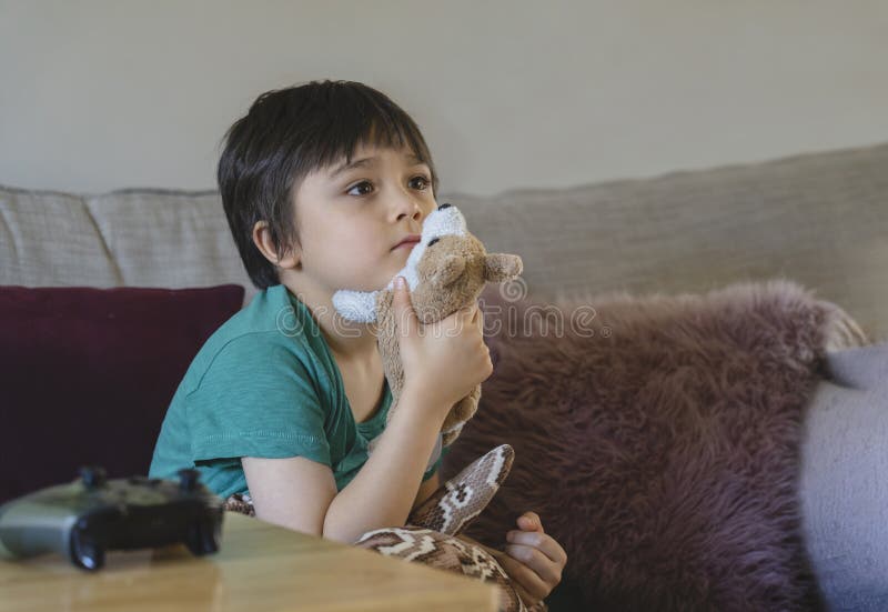 Emotional portrait of kid sitting on sofa playing his soft dog toy looking out. Child with curious face while watching cartoon on TV, Little boy stay at home relaxing in living room. Emotional portrait of kid sitting on sofa playing his soft dog toy looking out. Child with curious face while watching cartoon on TV, Little boy stay at home relaxing in living room