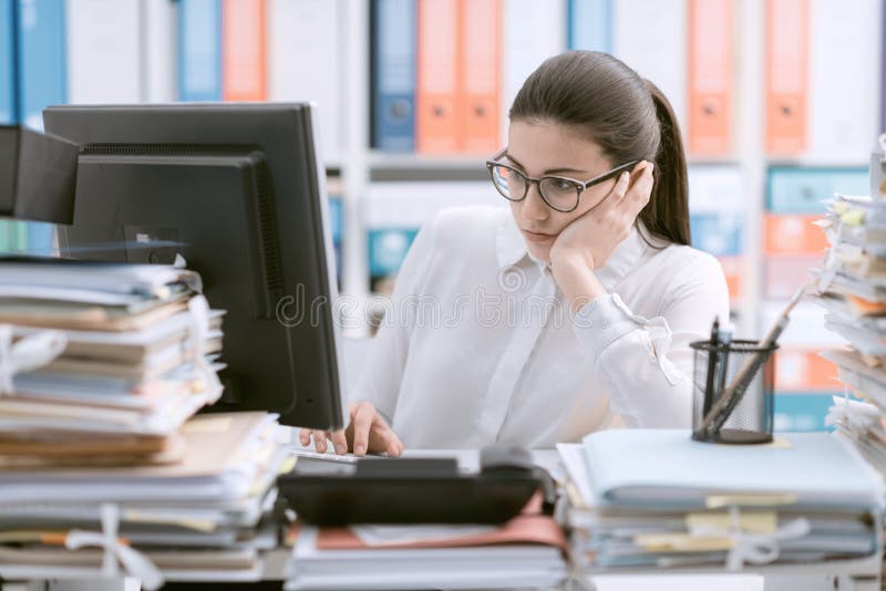 Young bored office worker sitting at desk and working, she is overloaded with paperwork. Young bored office worker sitting at desk and working, she is overloaded with paperwork