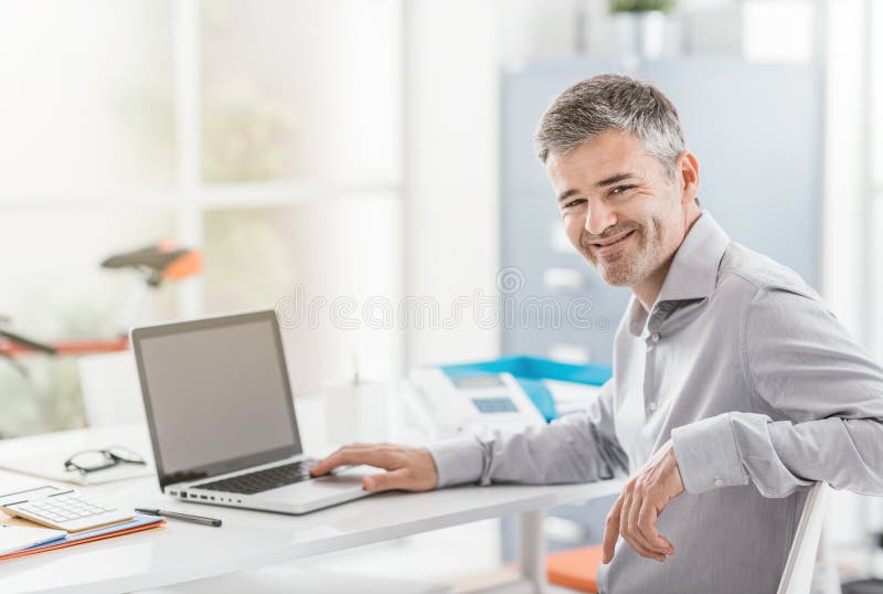 Employé de bureau sûr s'asseyant au bureau et travaillant avec un ordinateur portable, il sourit à l'appareil-photo photographie stock Employé de bureau sûr s'asseyant au bureau et travaillant avec un ordinateur portable, il sourit à l'appareil-photo photographie stock