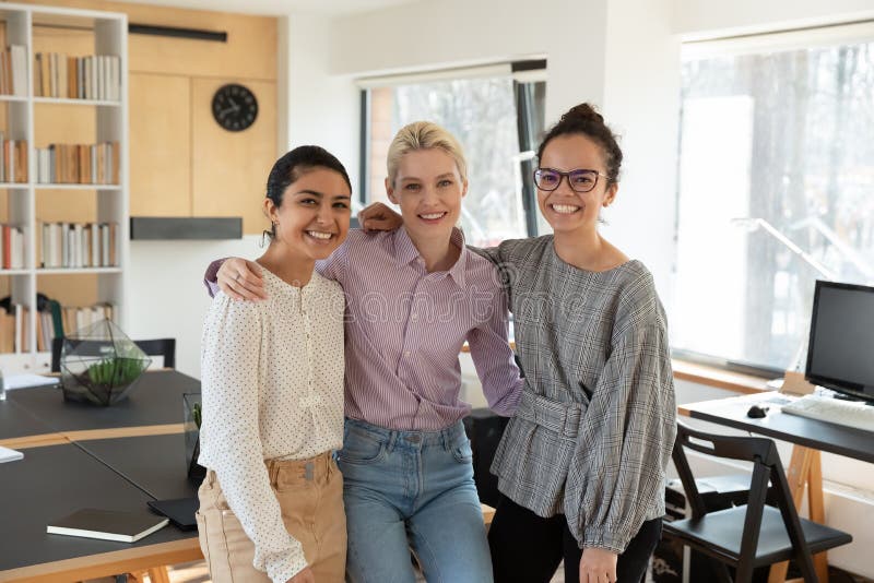 Portrait of smiling young european women hugging happy indian and african american female colleagues in creative office, friendly mixed race employees looking at camera, working relations concept. Portrait of smiling young european women hugging happy indian and african american female colleagues in creative office, friendly mixed race employees looking at camera, working relations concept