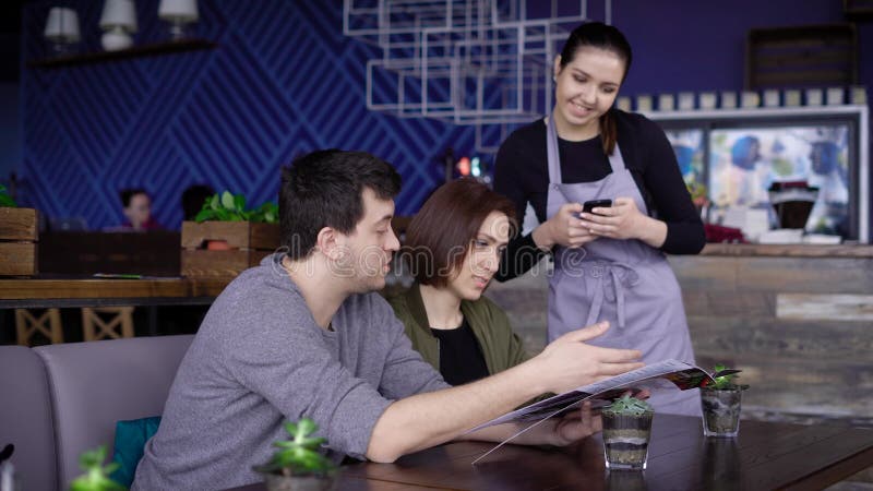 Empregada de mesa de sorriso nova que está no café perto da tabela de pares felizes e que põe sua ordem na tabuleta digital Homem