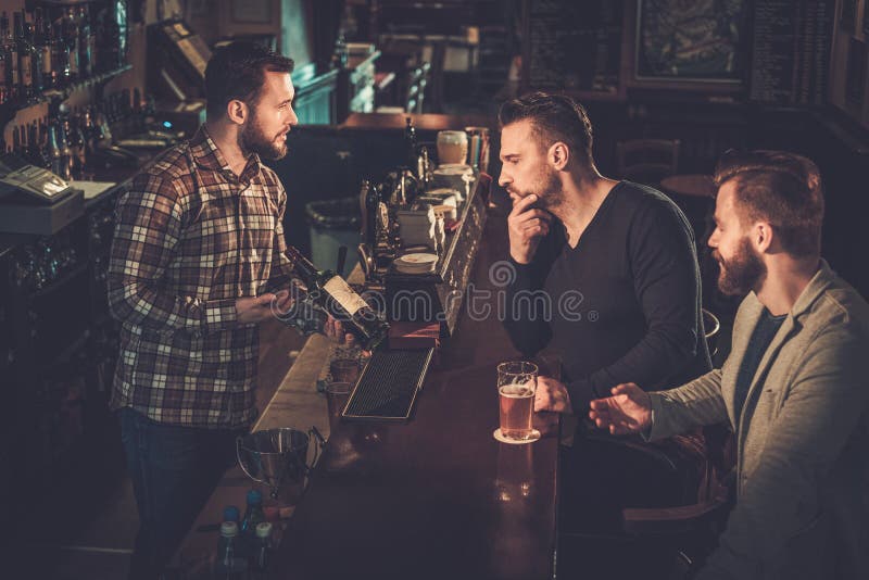 Empregado de bar polido que mostra uma garrafa de uísque aos clientes no contador da barra no bar foto de stock Empregado de bar polido que mostra uma garrafa de uísque aos clientes no contador da barra no bar foto de stock