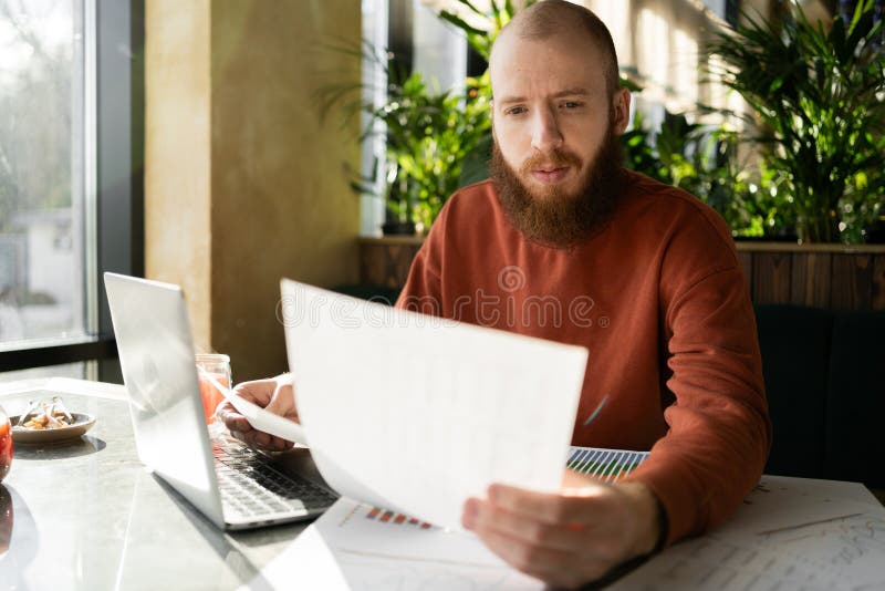 Emprendimiento. Hombre apuesto barbudo leyendo documento sentado con un portátil en la cafetería cerca de la ventana. Inicio de ne foto de archivo