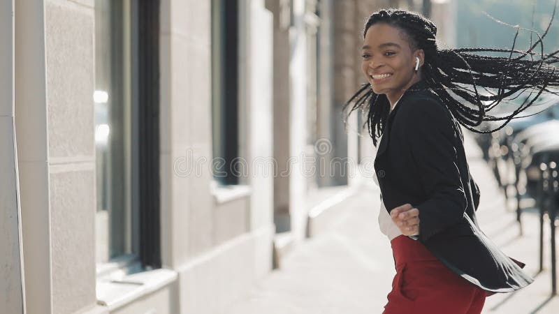 Empresaria afroamericana feliz joven en auriculares que camina la calle de la ciudad mientras que escucha la música en