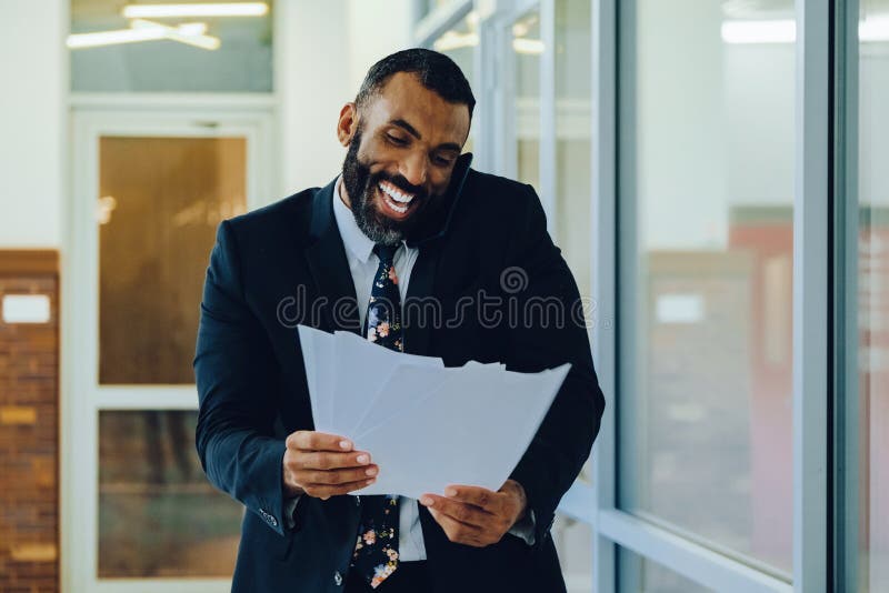 Empresario adulto medio barbudo hombre negro hombre de negocios usando traje portando papeles y hablando en un smartphone riendo imagen de archivo