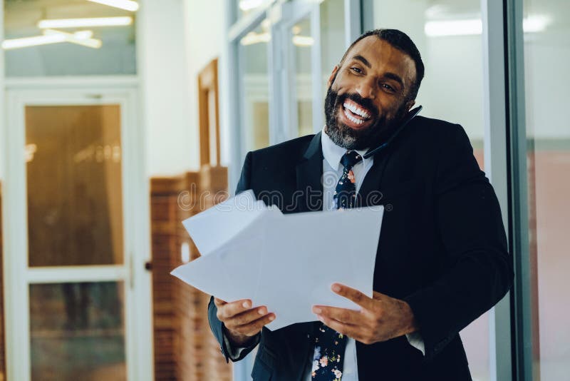 Empresario adulto medio barbudo hombre negro hombre de negocios usando traje portando papeles y hablando en un smartphone riendo imagen de archivo