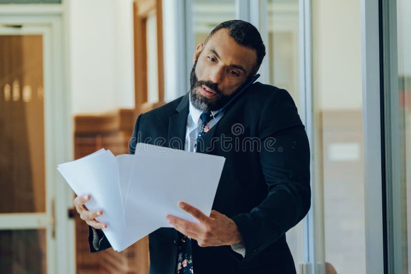 Empresario adulto medio barbudo hombre negro hombre de negocios vestido con traje portando papeles y hablando en smartphone estres fotografía de archivo