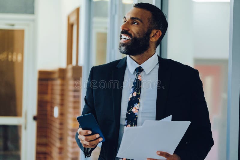 Empresario adulto medio barbudo hombre negro hombre de negocios vestido con traje portando papeles y smartphone caminando en el ca imagenes de archivo
