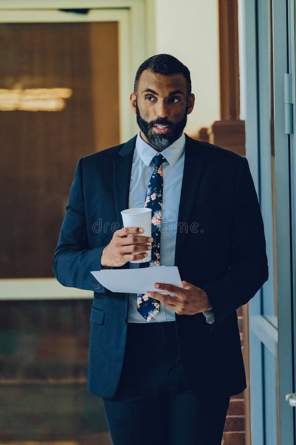 Empresario adulto medio barbudo hombre negro hombre de negocios vestido con traje sujetando una taza de café y papeles caminando e fotografía de archivo