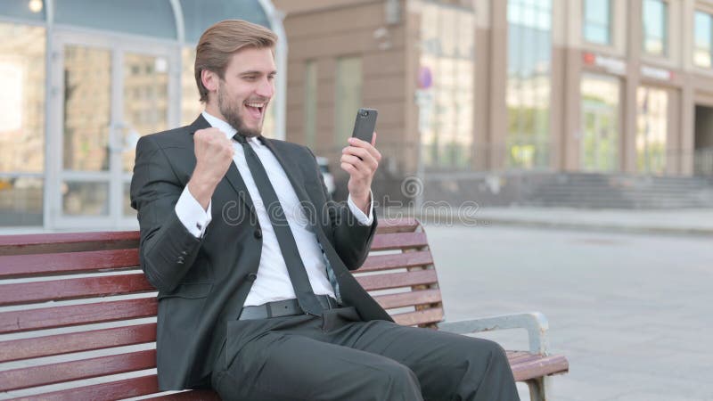 Empresario celebrando el éxito en línea en una sesión de teléfono inteligente al aire libre en el banco imagenes de archivo