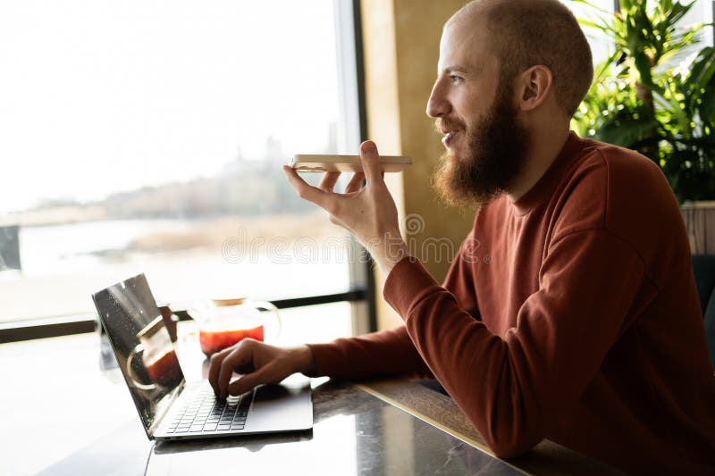 Empresario con suéter caliente usando un smartphone mientras trabaja en un portátil en un acogedor café verde fotos de archivo