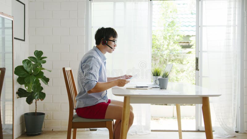 Empresario de asia vestido con camisa y pantalones cortos usa laptop para hablar con colegas en videollamadas mientras trabaja des almacen de video