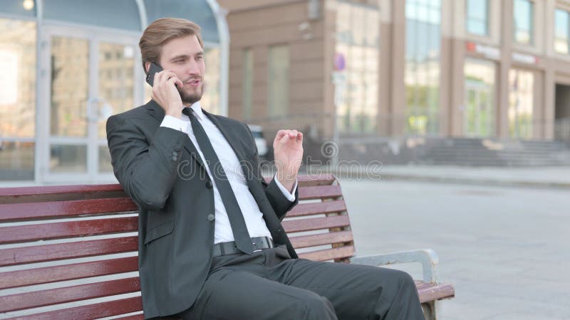 Empresario hablando por teléfono mientras está sentado al aire libre en un banco imagen de archivo libre de regalías