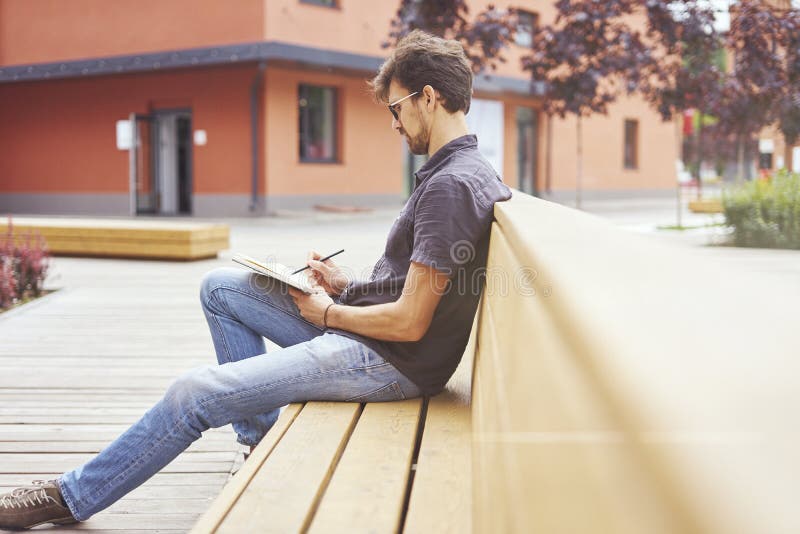 Empresario joven que trabaja en el parque afuera en banco de madera Vidrios que llevan del hombre hermoso usando el libro, escrib foto de archivo Empresario joven que trabaja en el parque afuera en banco de madera Vidrios que llevan del hombre hermoso usando el libro, escrib foto de archivo