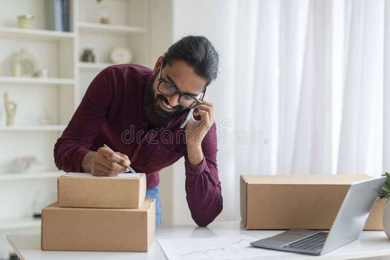 Smiling indian entrepreneur managing logistics during phone call while labeling packages, young man multitasking in his home office, standing at desk with laptop and business charts, copy space. Smiling indian entrepreneur managing logistics during phone call while labeling packages, young man multitasking in his home office, standing at desk with laptop and business charts, copy space