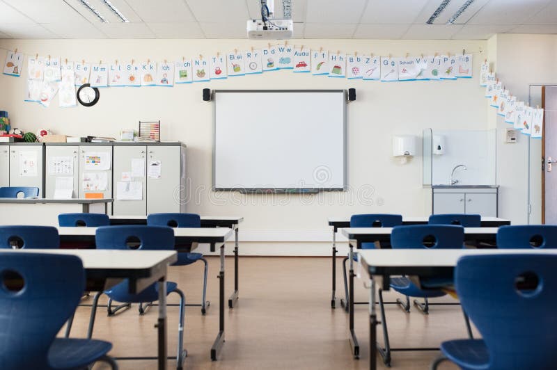 School desk and chairs in empty modern classroom. Empty class room with white board and projector in elementary school. Primary classroom with smartboard and alphabet on wall. School desk and chairs in empty modern classroom. Empty class room with white board and projector in elementary school. Primary classroom with smartboard and alphabet on wall.