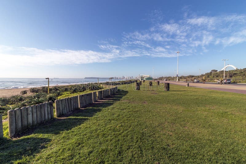 Empty Gass Verge Beachfront against City Skyline. Empty grass verge lined paved promenade dune vegetation and sea against distant Durban city blue cloudy skyline royalty free stock photography Empty Gass Verge Beachfront against City Skyline. Empty grass verge lined paved promenade dune vegetation and sea against distant Durban city blue cloudy skyline royalty free stock photography