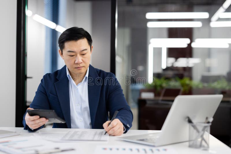 Focused Asian businessman reviewing financial reports while working in a modern office space. Concept of professionalism and diligence. Focused Asian businessman reviewing financial reports while working in a modern office space. Concept of professionalism and diligence.