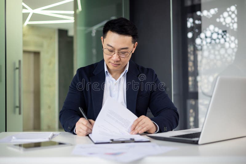 Focused Asian businessman working diligently on business documents, sitting at a white table in a well-lit contemporary office environment. Focused Asian businessman working diligently on business documents, sitting at a white table in a well-lit contemporary office environment.