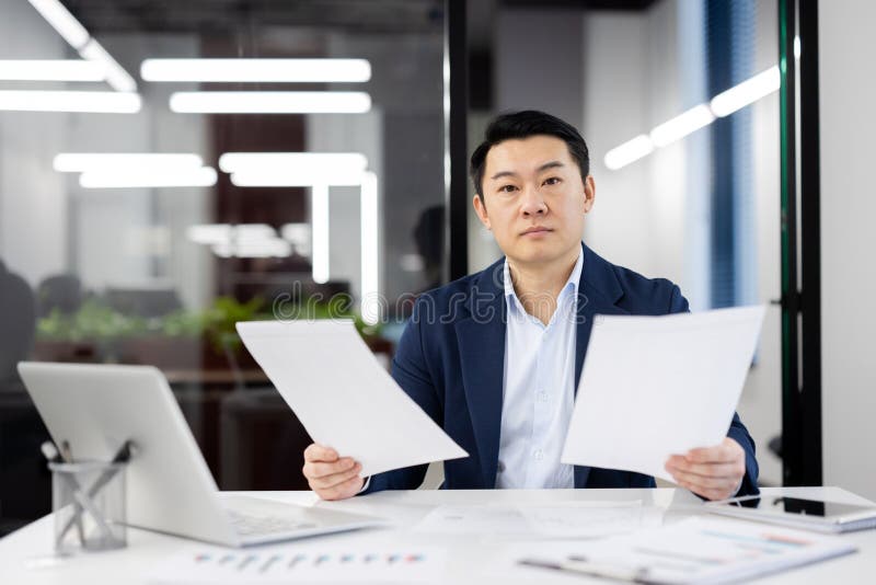 Focused Asian businessman analyzing paperwork while sitting at his desk in a contemporary office setting with laptop and plants in the background. Focused Asian businessman analyzing paperwork while sitting at his desk in a contemporary office setting with laptop and plants in the background.