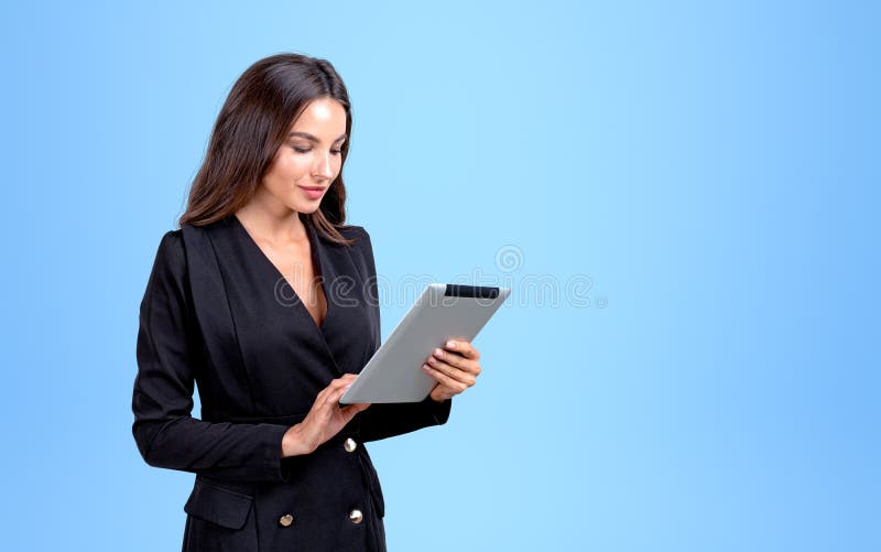 Attractive businesswoman wearing formal wear is holding tablet device standing in front of blue wall in background. Concept of imagination and inspiration for creative ideas, gadgets and communication. Attractive businesswoman wearing formal wear is holding tablet device standing in front of blue wall in background. Concept of imagination and inspiration for creative ideas, gadgets and communication